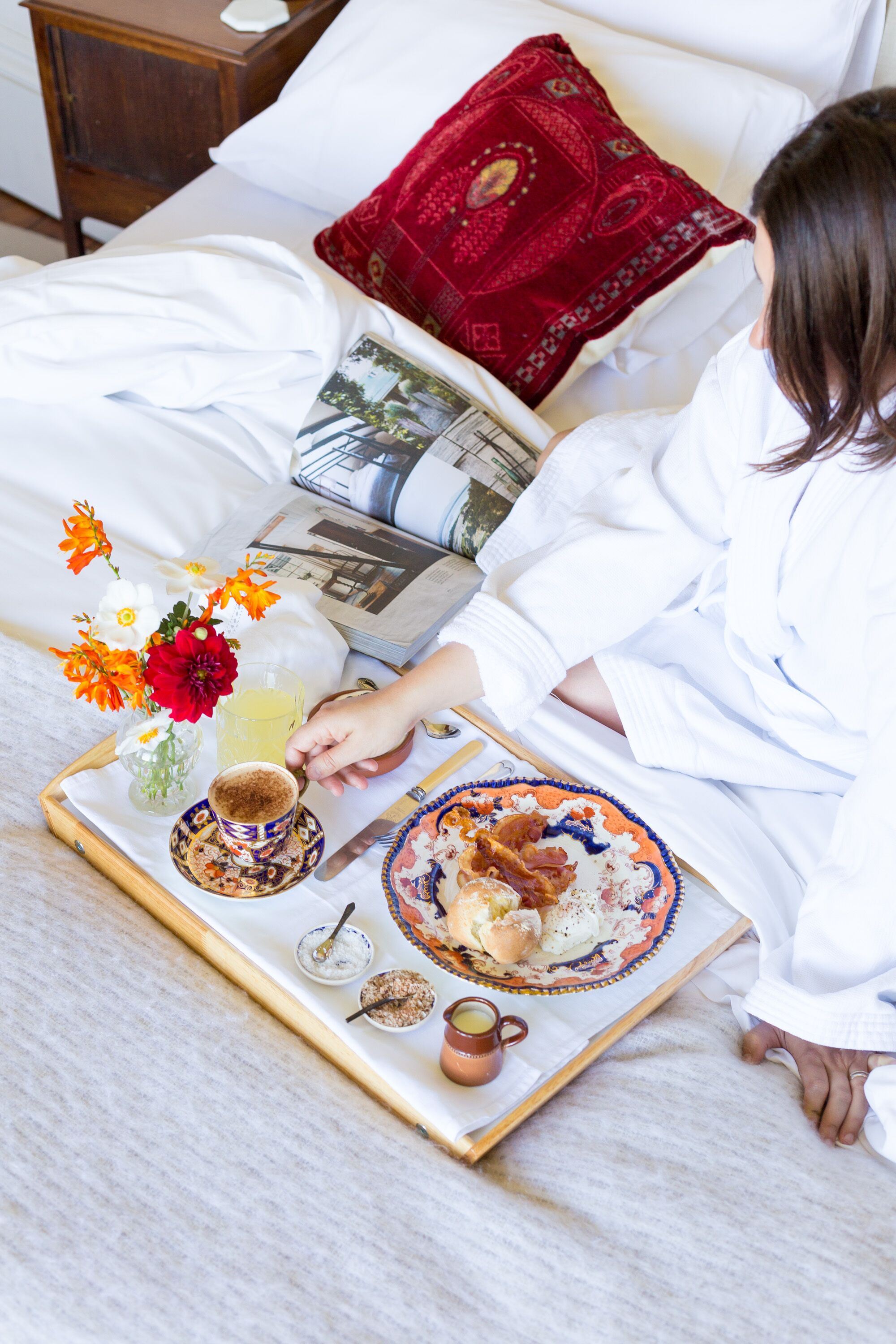 Uma pessoa vestindo um robe branco sentada em uma cama com uma bandeja de comida, um livro e um travesseiro vermelho.