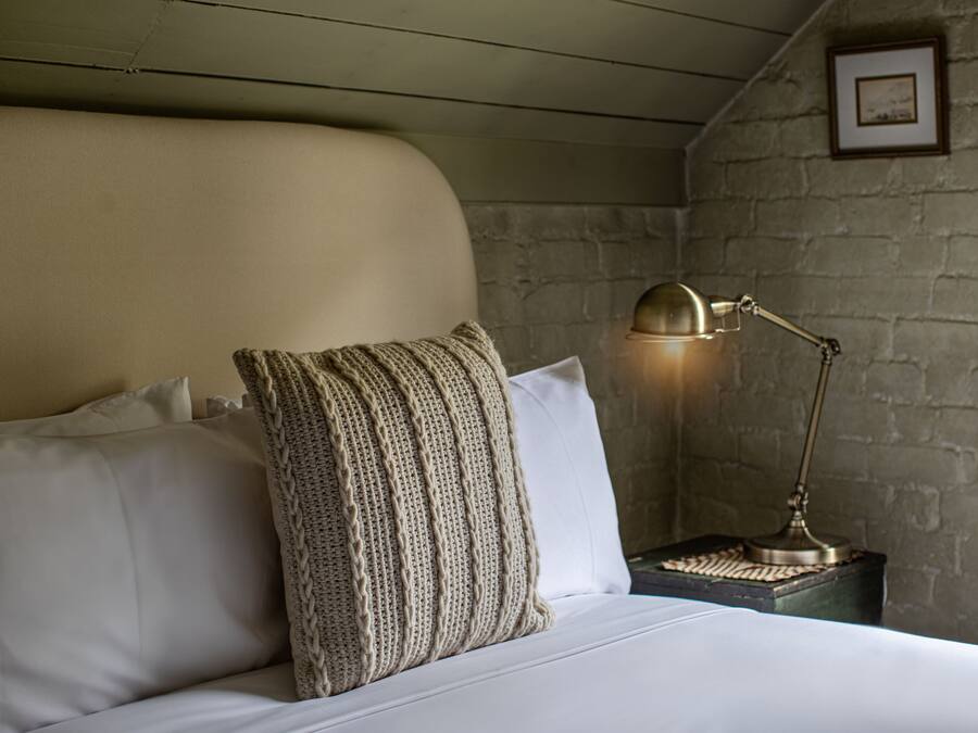 Un lit bien fait, avec du linge de lit blanc et un coussin décoratif à motifs, une lampe de bureau en laiton et un tableau encadré accroché au mur.