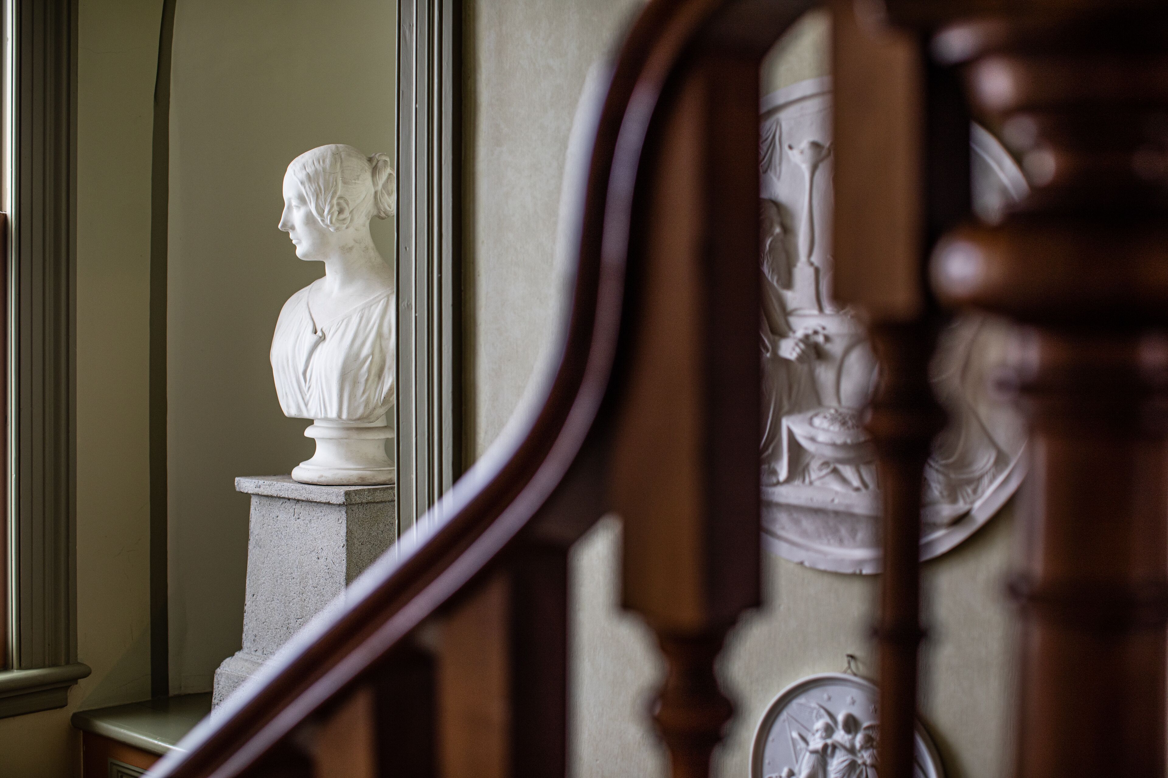 Un busto de mármol sobre un pedestal en una sala de arquitectura clásica, que incluye una escalera de madera y un muro con relieves decorativos.