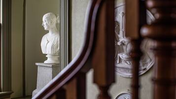 Un busto de mármol sobre un pedestal en una sala de arquitectura clásica, que incluye una escalera de madera y un muro con relieves decorativos.