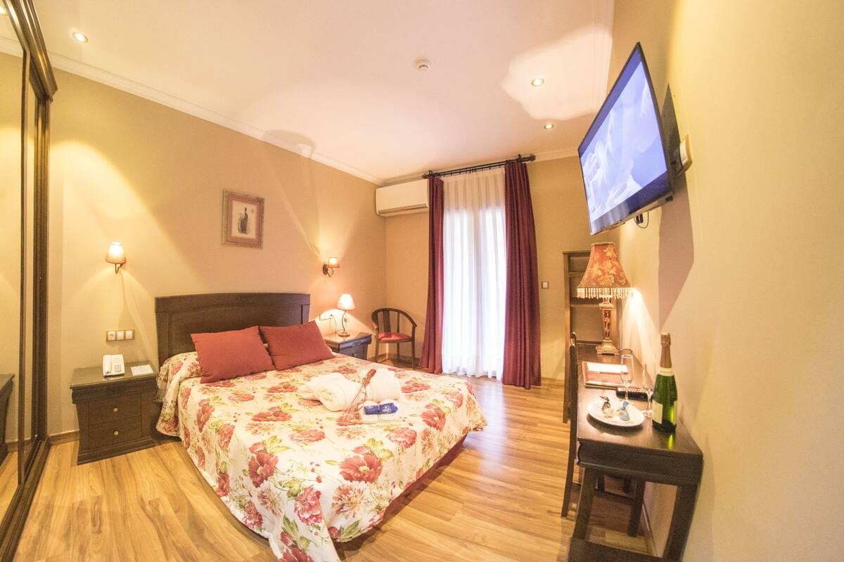 A hotel room with a bed, bedside tables, a TV, a desk with a chair, and a dining table set for two.
