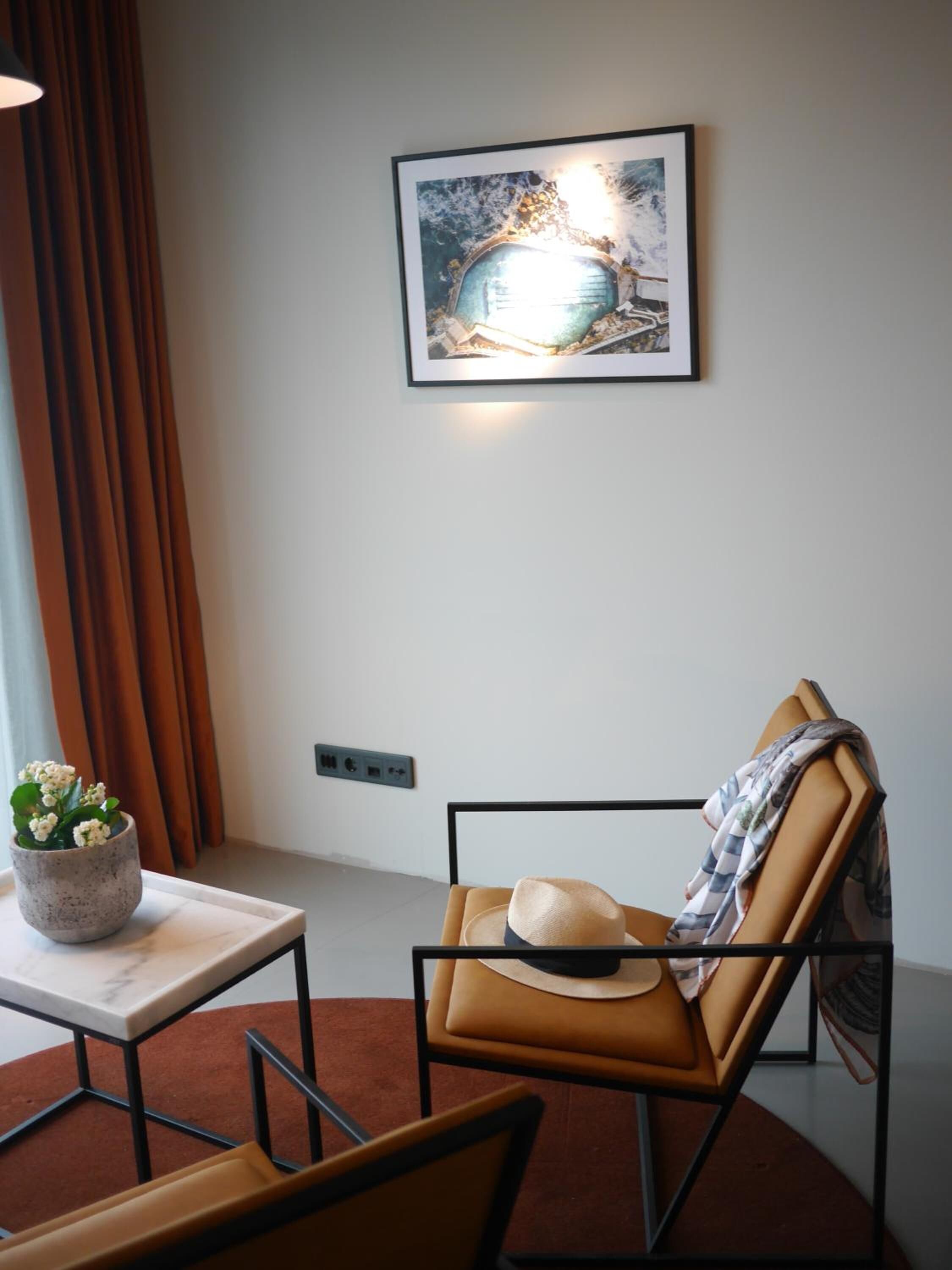 A modern room with a framed painting, a small table with a potted plant, and a leather chair with a hat.