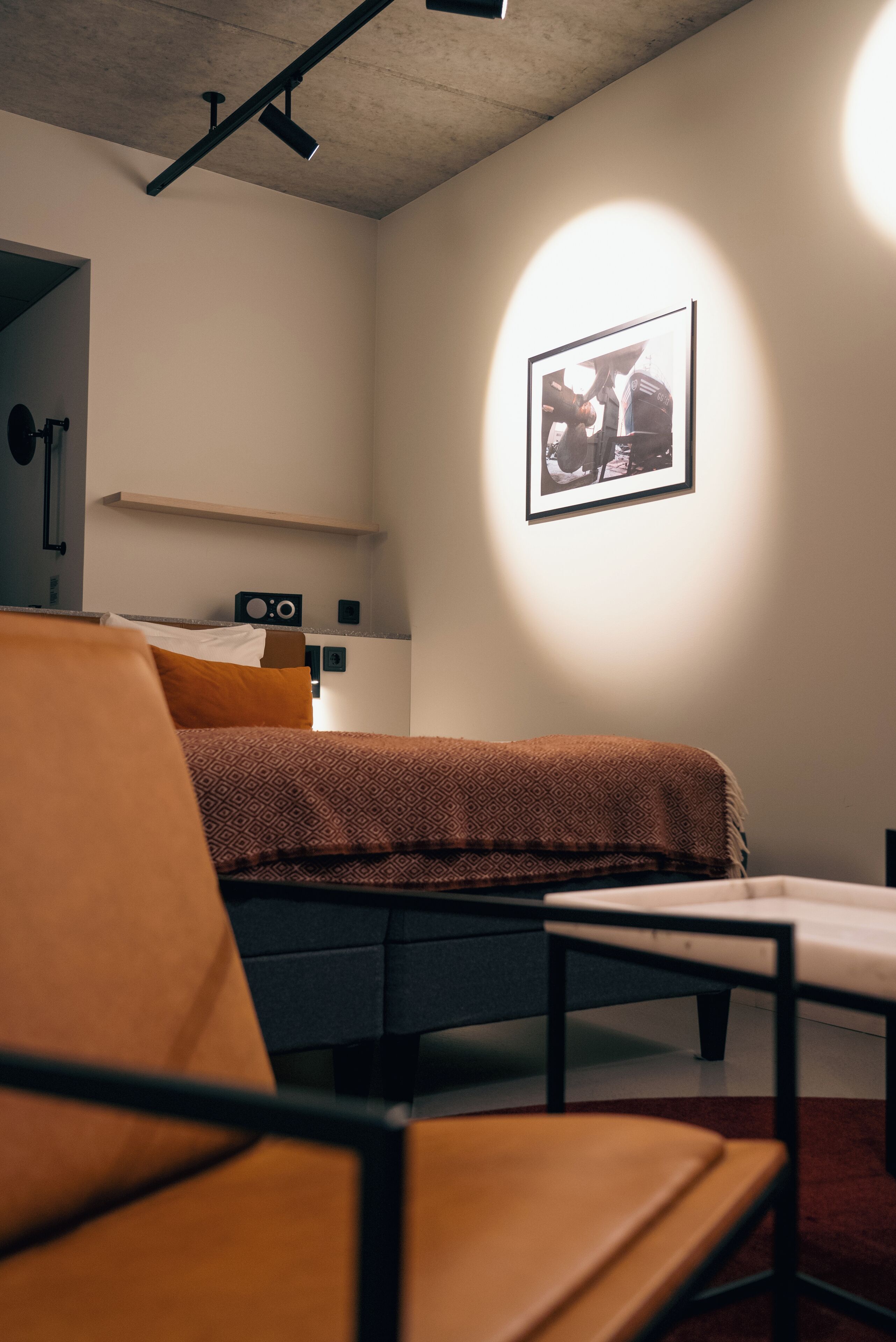 A modern hotel room with a bed, a chair, a desk, and a framed picture on the wall.