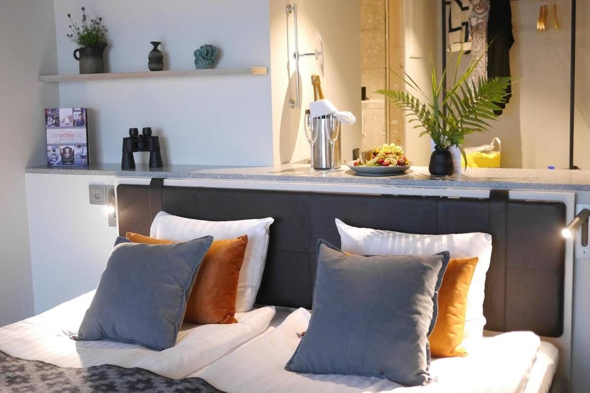 A neatly arranged bedroom with a bed, pillows, a bedside table with a lamp, and a shelf with decorative items.