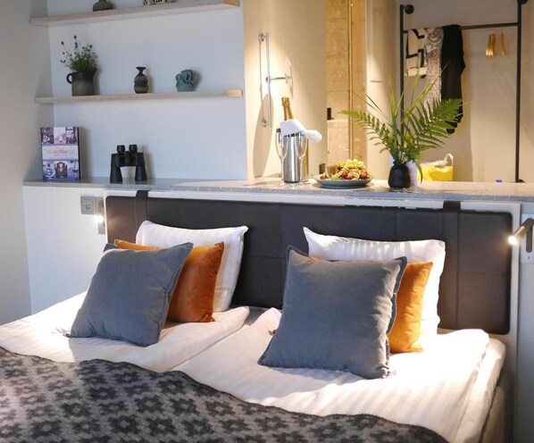 Une chambre à coucher bien rangée, avec un lit, des oreillers, une table de chevet avec une lampe et une étagère ornée d’objets décoratifs.