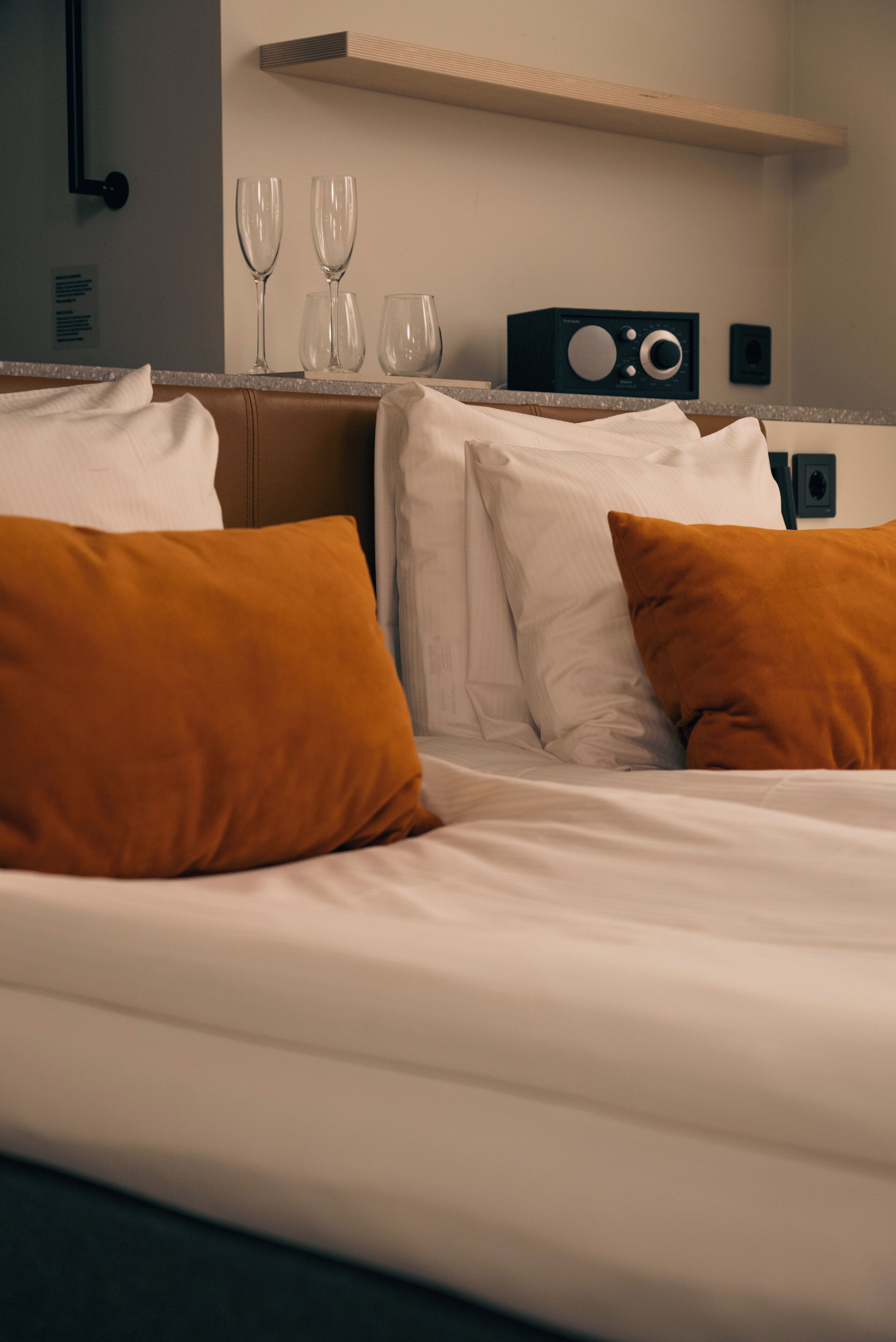 Ett hotellrum med en säng, vita och orange kuddar, en träfärgad sänggavel och ett litet bord med vinglas och en radio.