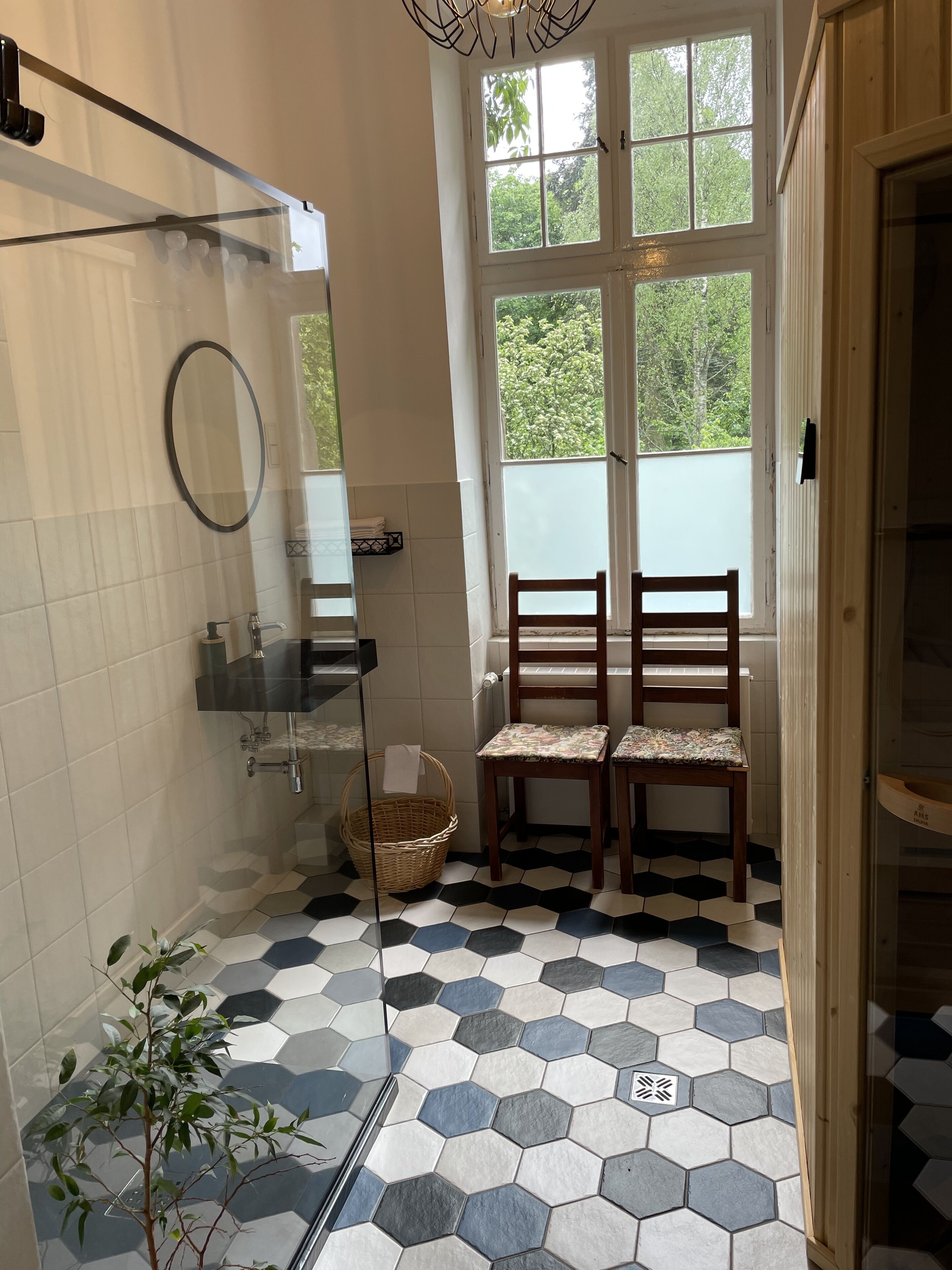 A bathroom with a hexagonal tile floor, a glass shower enclosure, a wooden chair, and a large window with a view of greenery.