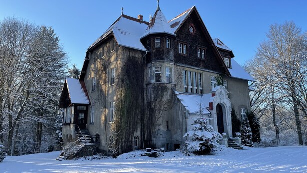 Ein großes, historisches Gebäude mit einem steilen Dach und zahlreichen Fenstern, umgeben von schneebedeckten Bäumen.