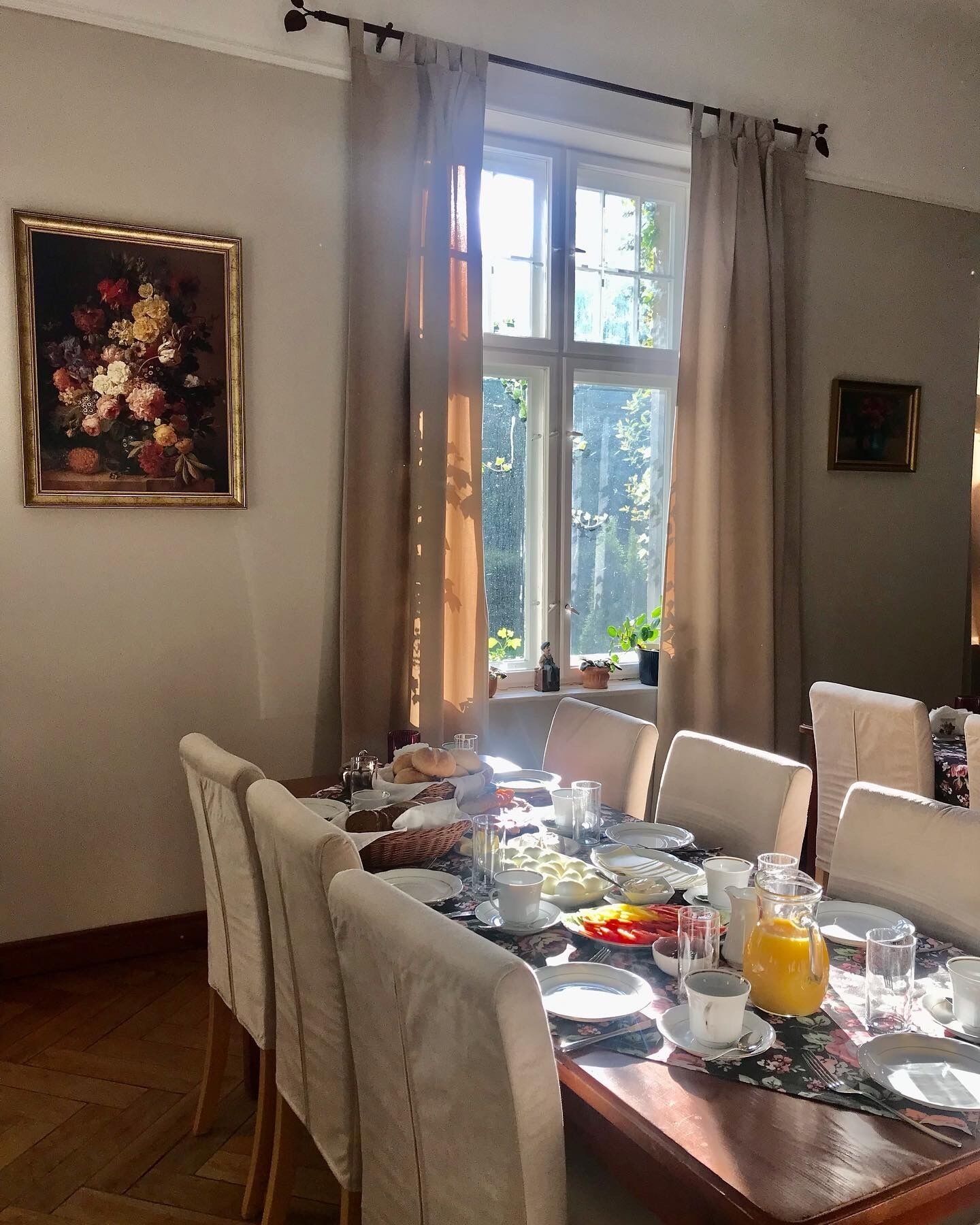 A dining area with a table set for breakfast, a painting on the wall, and a window with curtains.