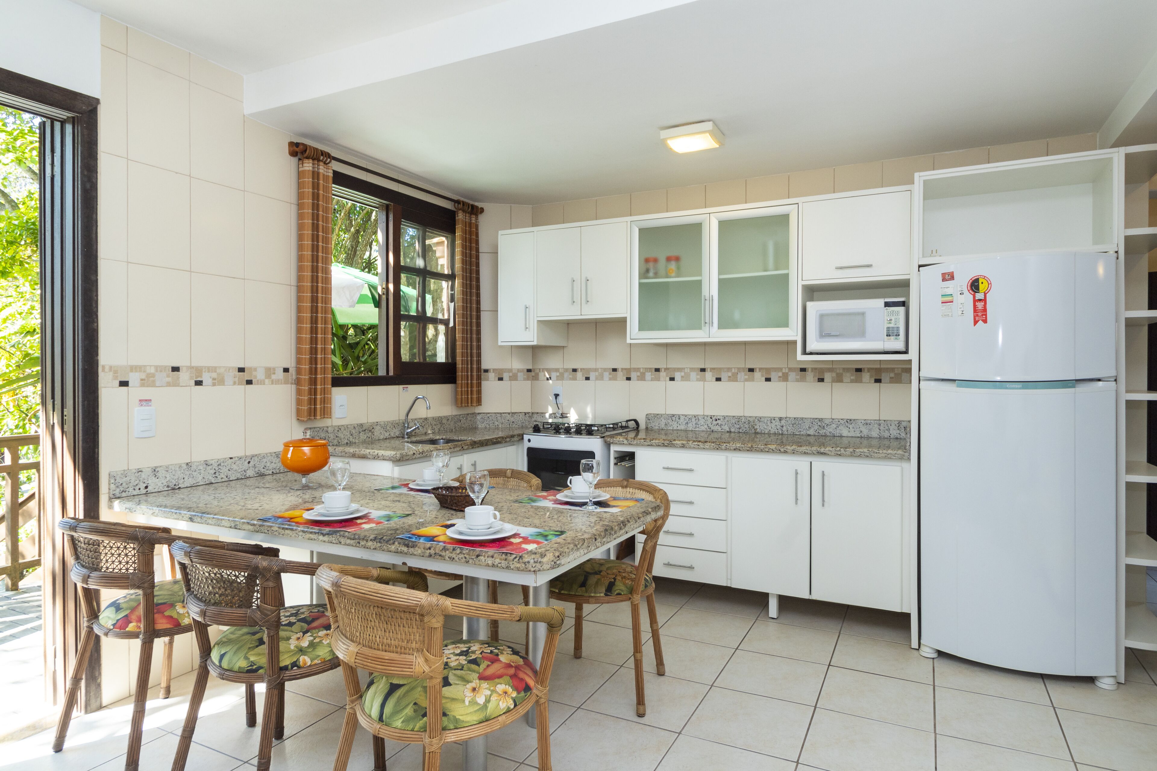 Une cuisine moderne avec des meubles blancs, un four à micro-ondes, un réfrigérateur et un coin repas agrémenté de chaises et d’une table en osier, le tout disposé pour accueillir quatre personnes.