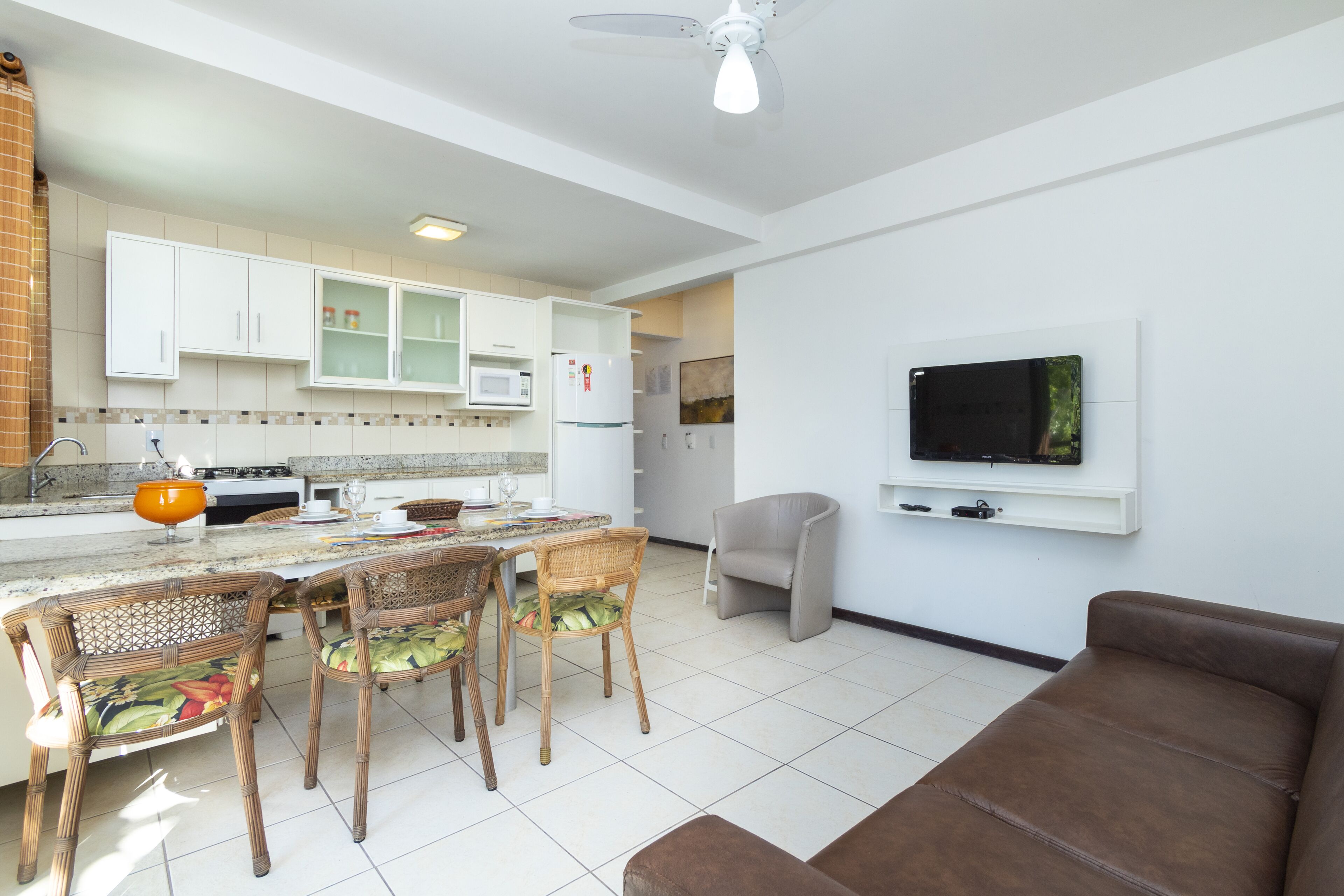 Eine moderne Küche mit weißen Schränken, ein Essbereich mit Korbstühlen, ein braunes Sofa und ein Flachbildfernseher an der Wand.