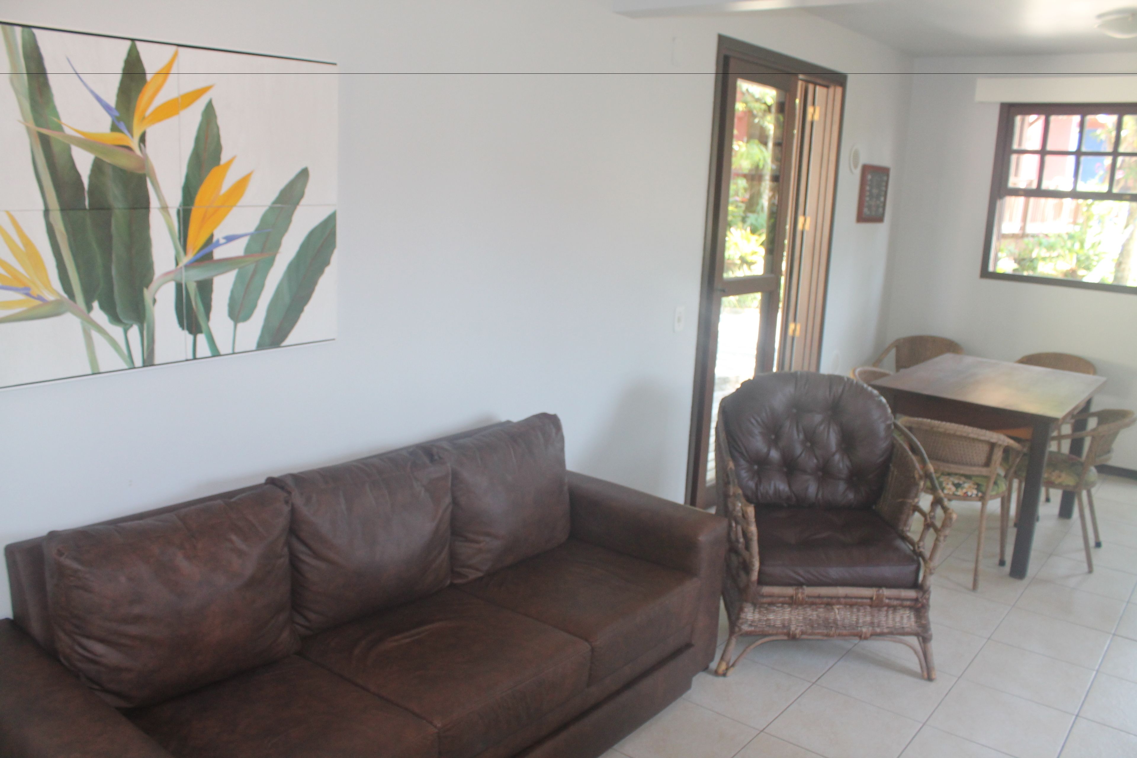 A brown leather sofa, wicker chair, dining table, and chairs in a room with white walls and a window.