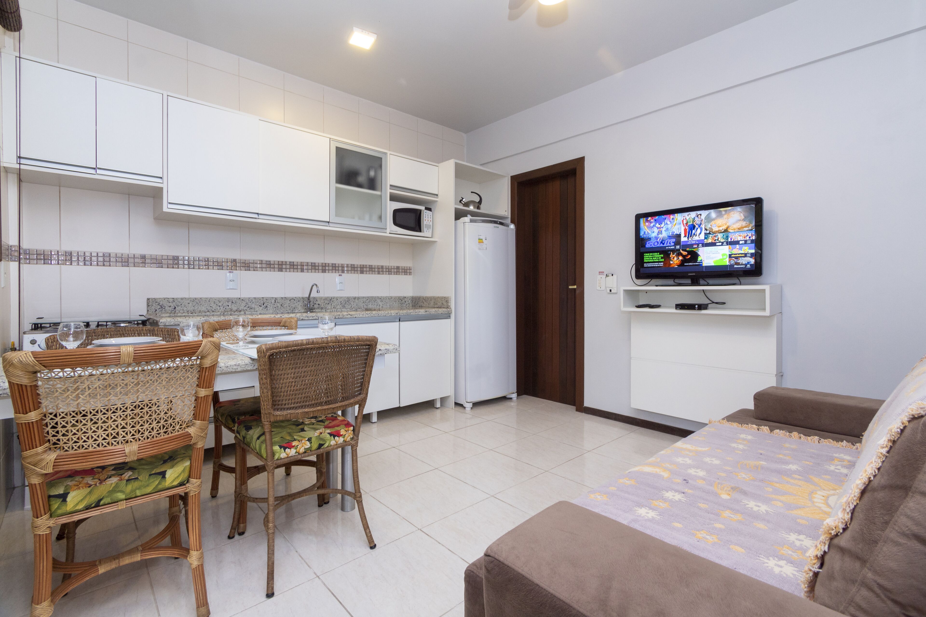 Una cocina moderna con armarios blancos, un microondas y un refrigerador. Hay una zona de comedor con sillas de mimbre y una mesa dispuesta para dos personas. Un televisor está montado en la pared sobre un sofá.