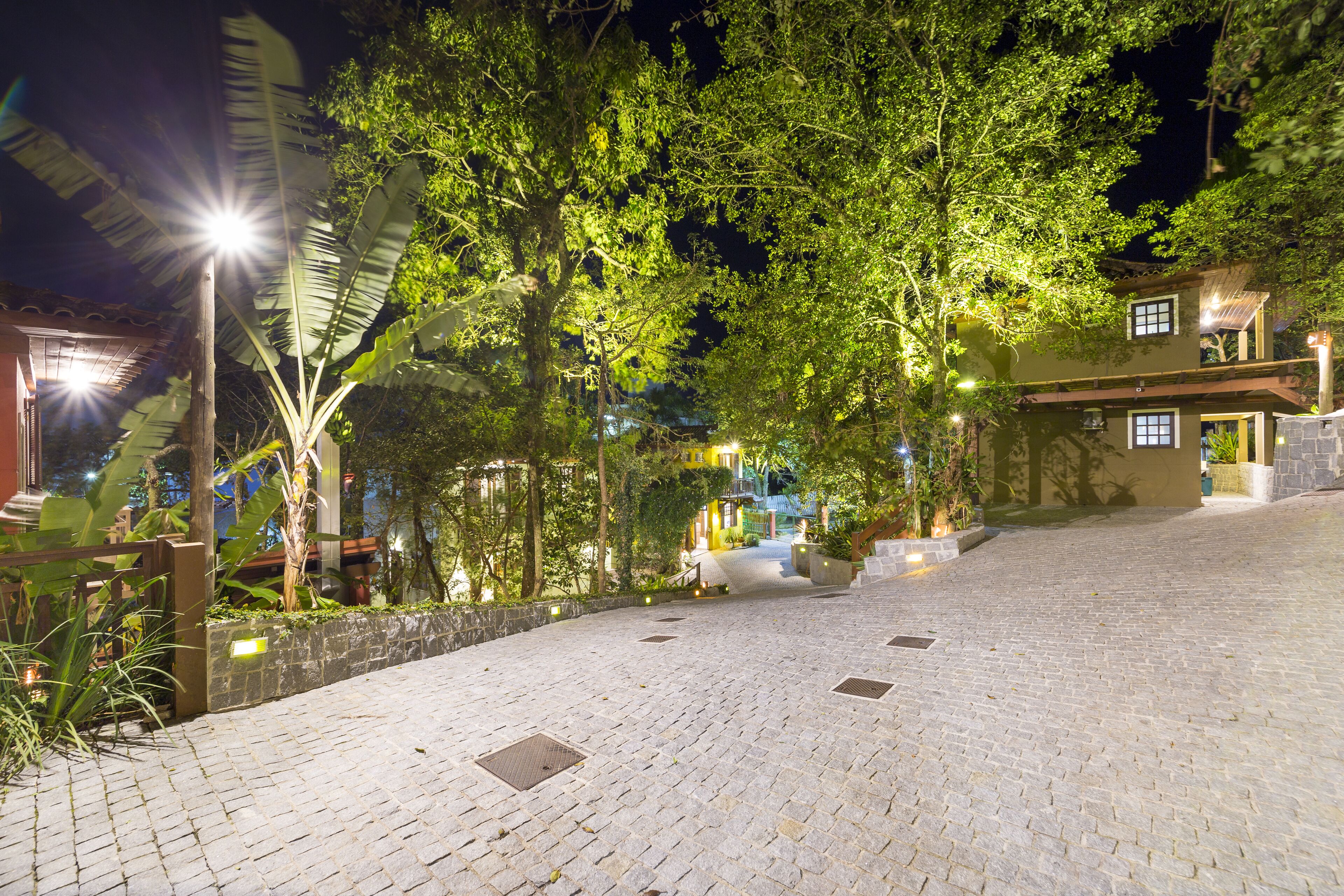 A paved pathway leading to a building with a sloped roof, surrounded by lush greenery and illuminated by streetlights.