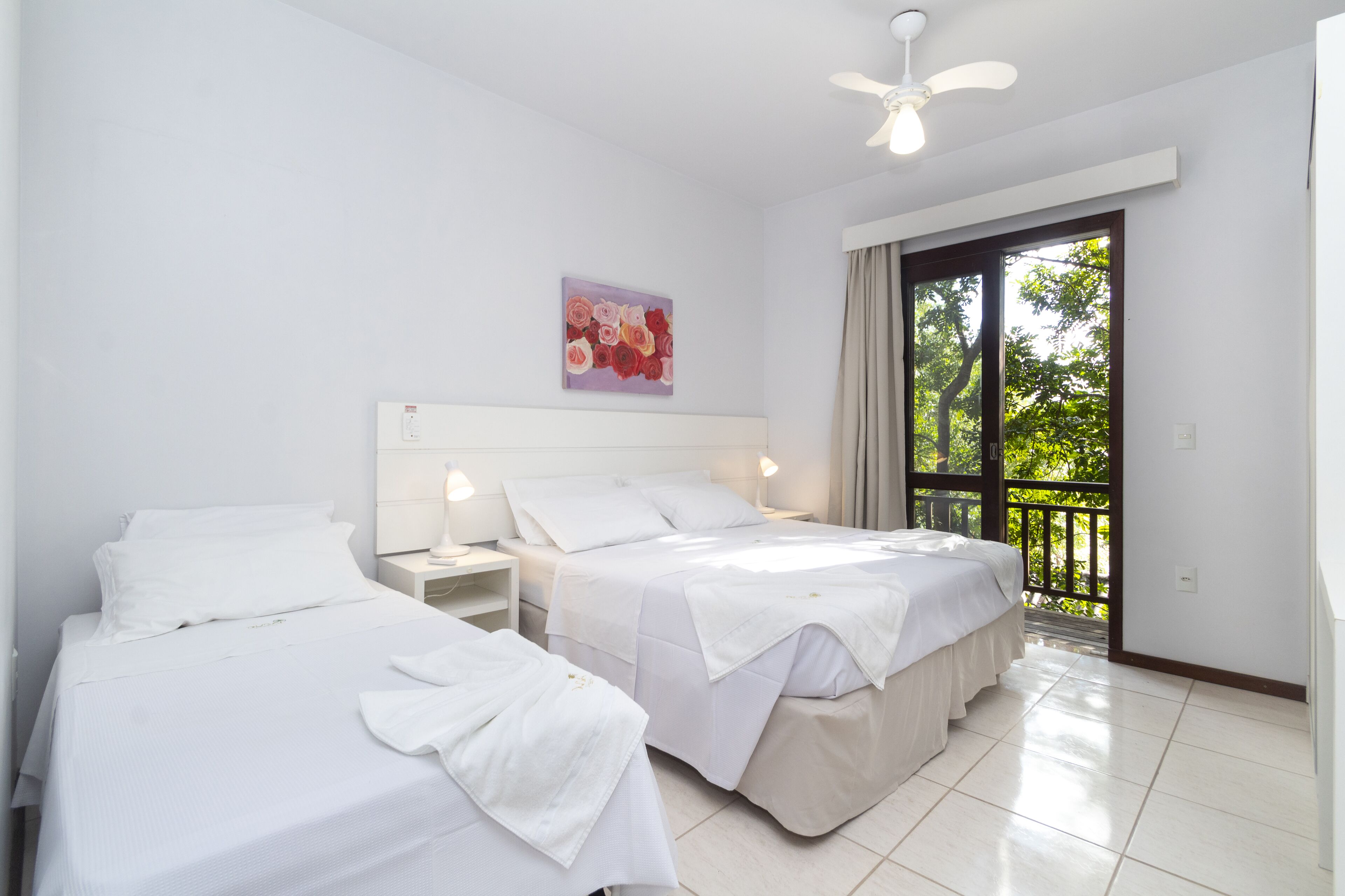 A hotel room with two beds, a ceiling fan, a painting, and a balcony with a view of trees.