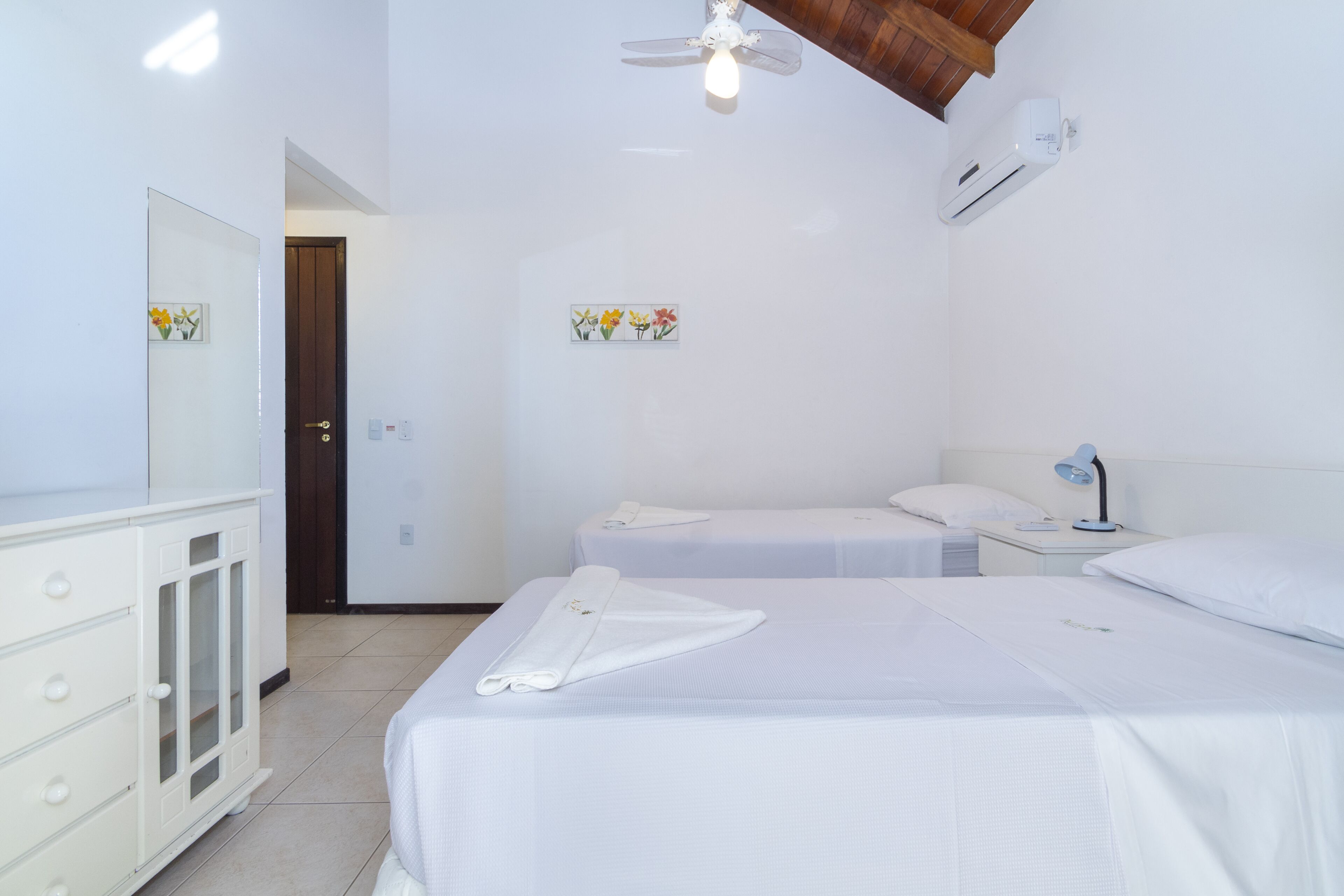 Une chambre propre et bien éclairée, avec deux lits, une armoire, un ventilateur de plafond et un climatiseur fixé au mur.