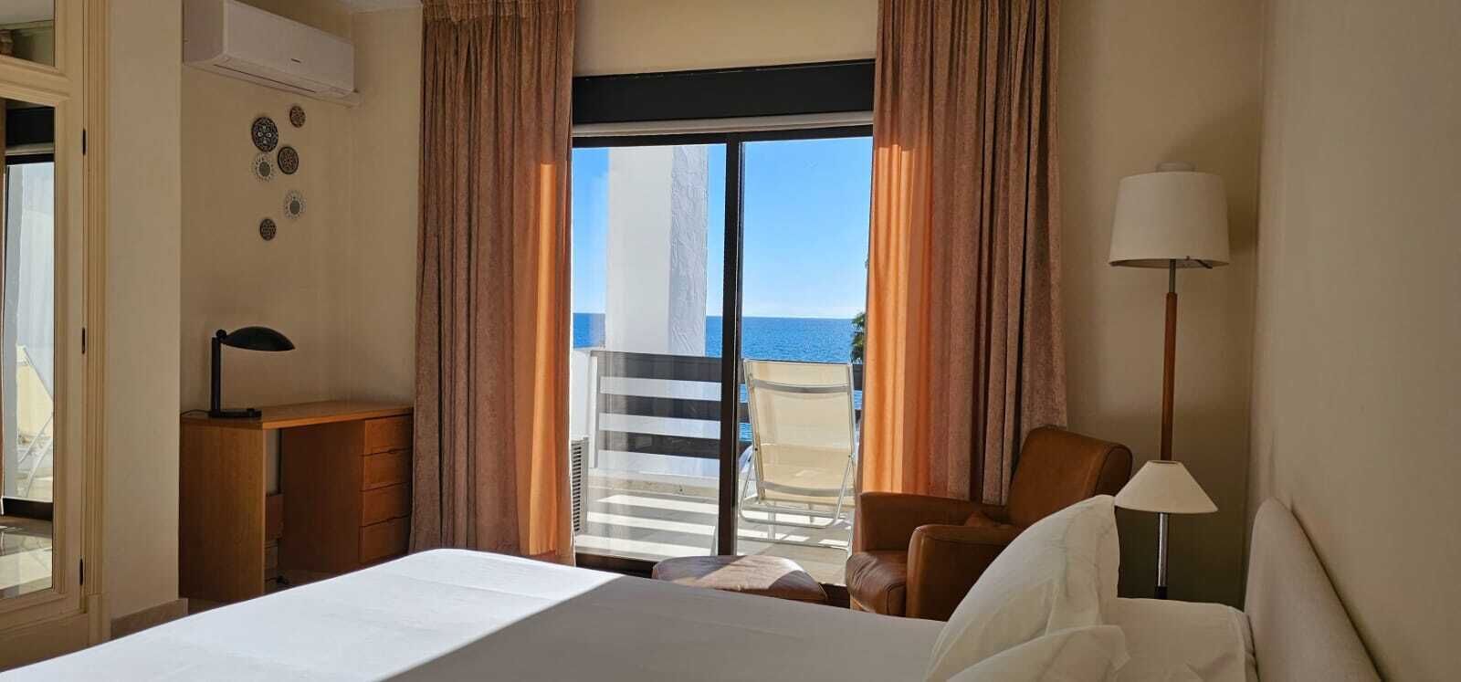 A hotel room with a bed, a desk, a chair, and a view of the sea through large windows.