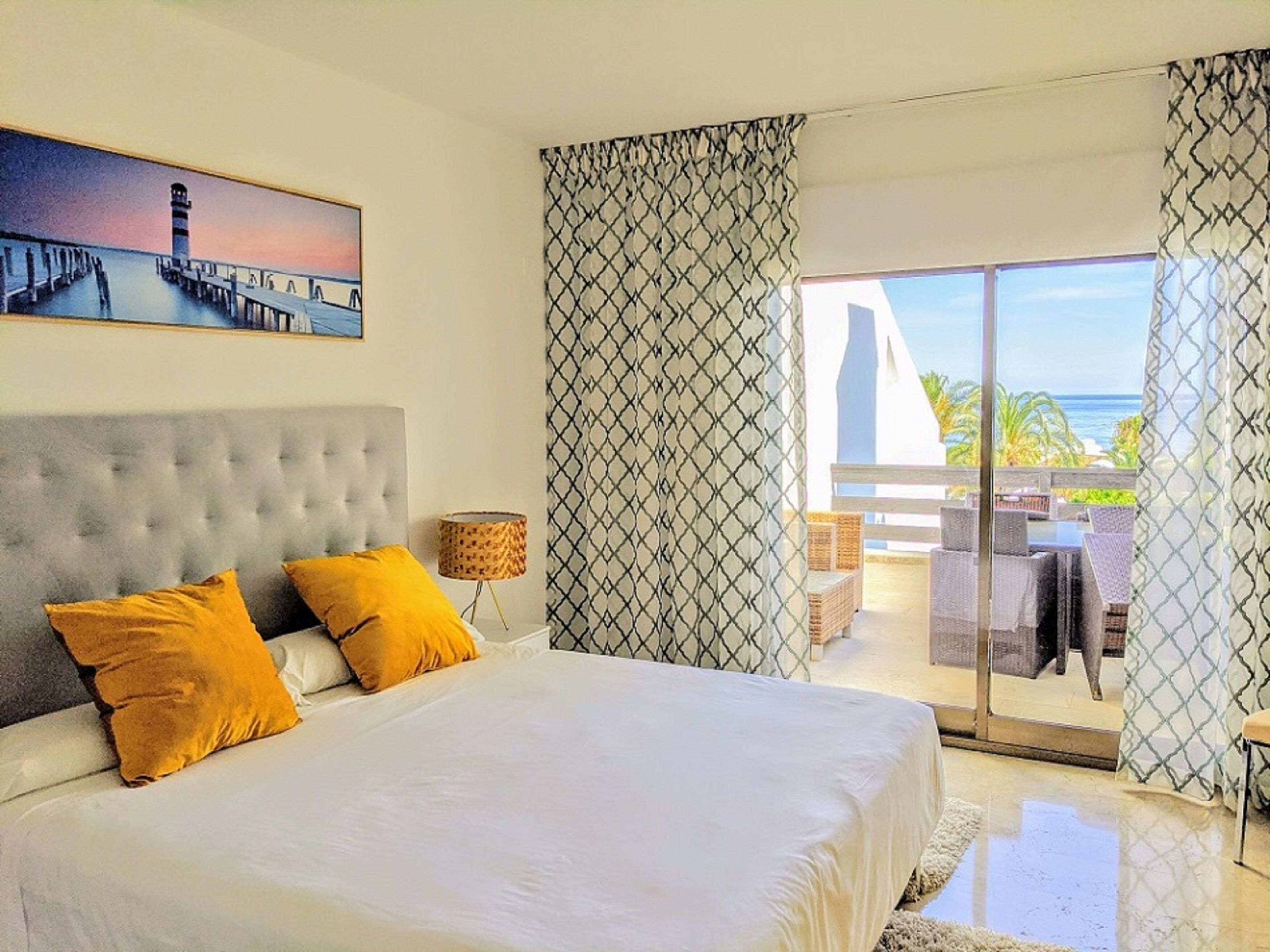 A bedroom with a large bed, two yellow pillows, a grey headboard, a painting of a pier, and a balcony with outdoor furniture.
