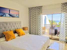 Slaapkamer met een groot bed, twee gele kussens, een grijs hoofdeinde, een schilderij van een pier en een balkon met tuinmeubilair.