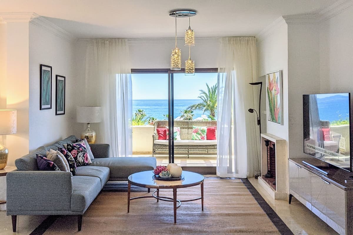 A living room with a sofa, a coffee table, a TV, and a view of the ocean.