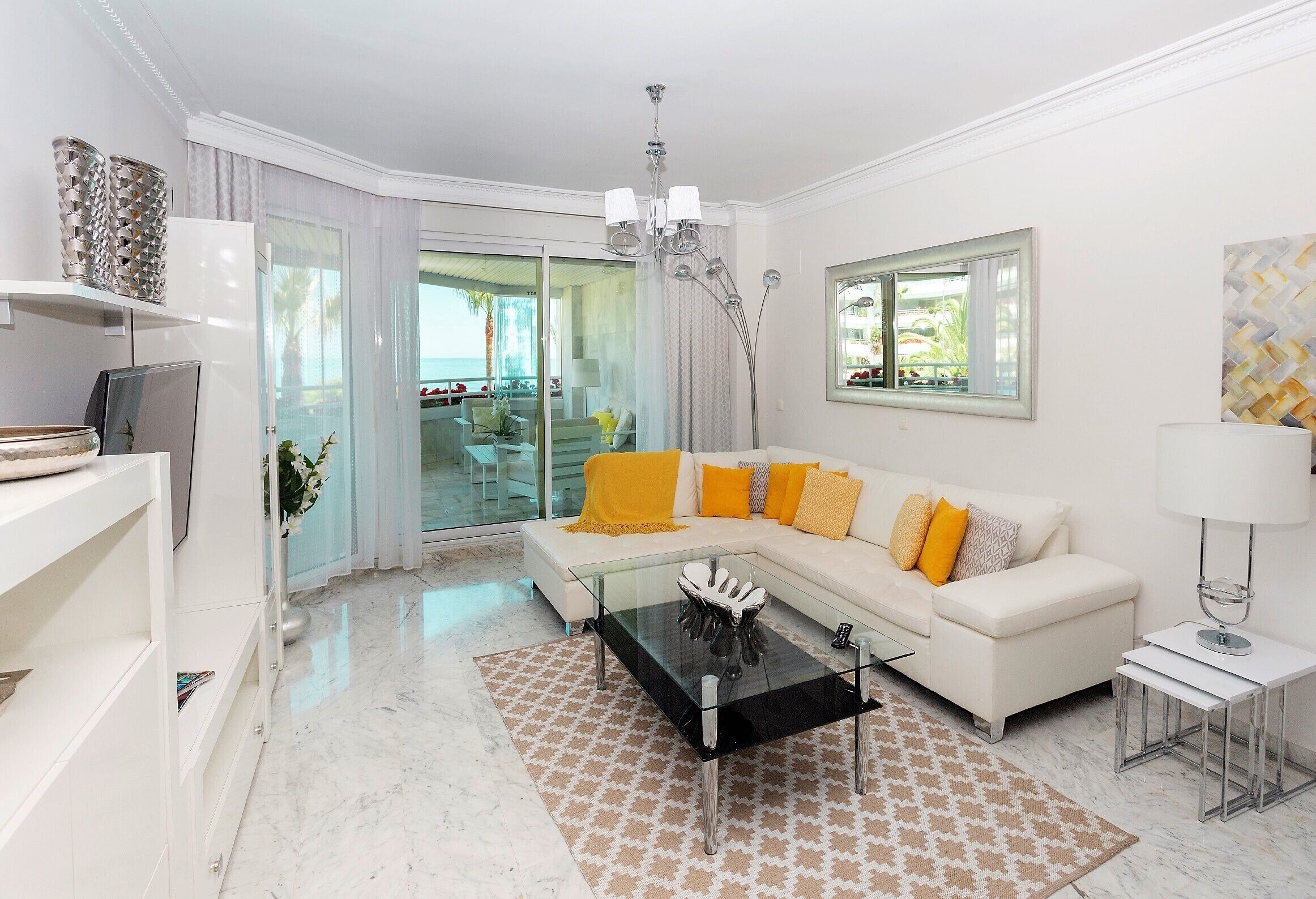 Una sala moderna con una mesa de centro de vidrio, un sofá blanco con cojines amarillos, una mesita auxiliar blanca y vistas al exterior a través de un ventanal.