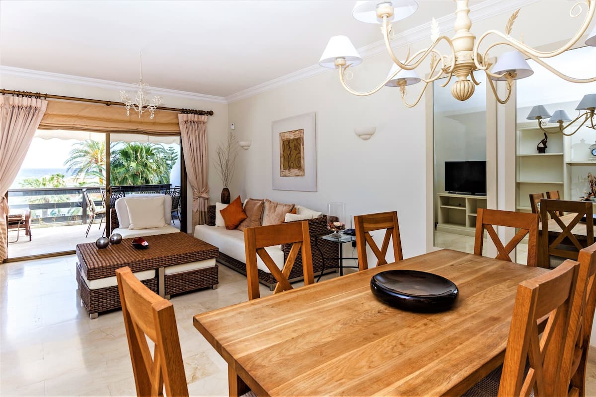 A spacious living room with a wooden dining table, comfortable seating, a television, and a balcony with a view of palm trees.