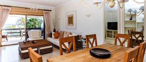 A spacious living room with a wooden dining table, comfortable seating, a television, and a balcony with a view of palm trees.