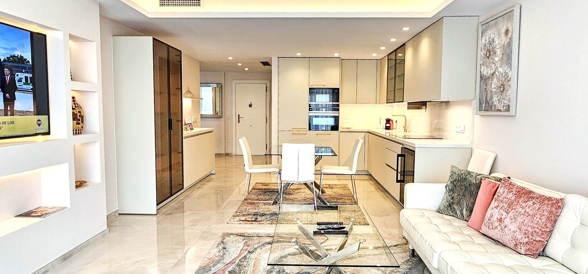 A modern living room with a white sofa, a coffee table, and a TV. There is a kitchen area with cabinets and a dining table with chairs.