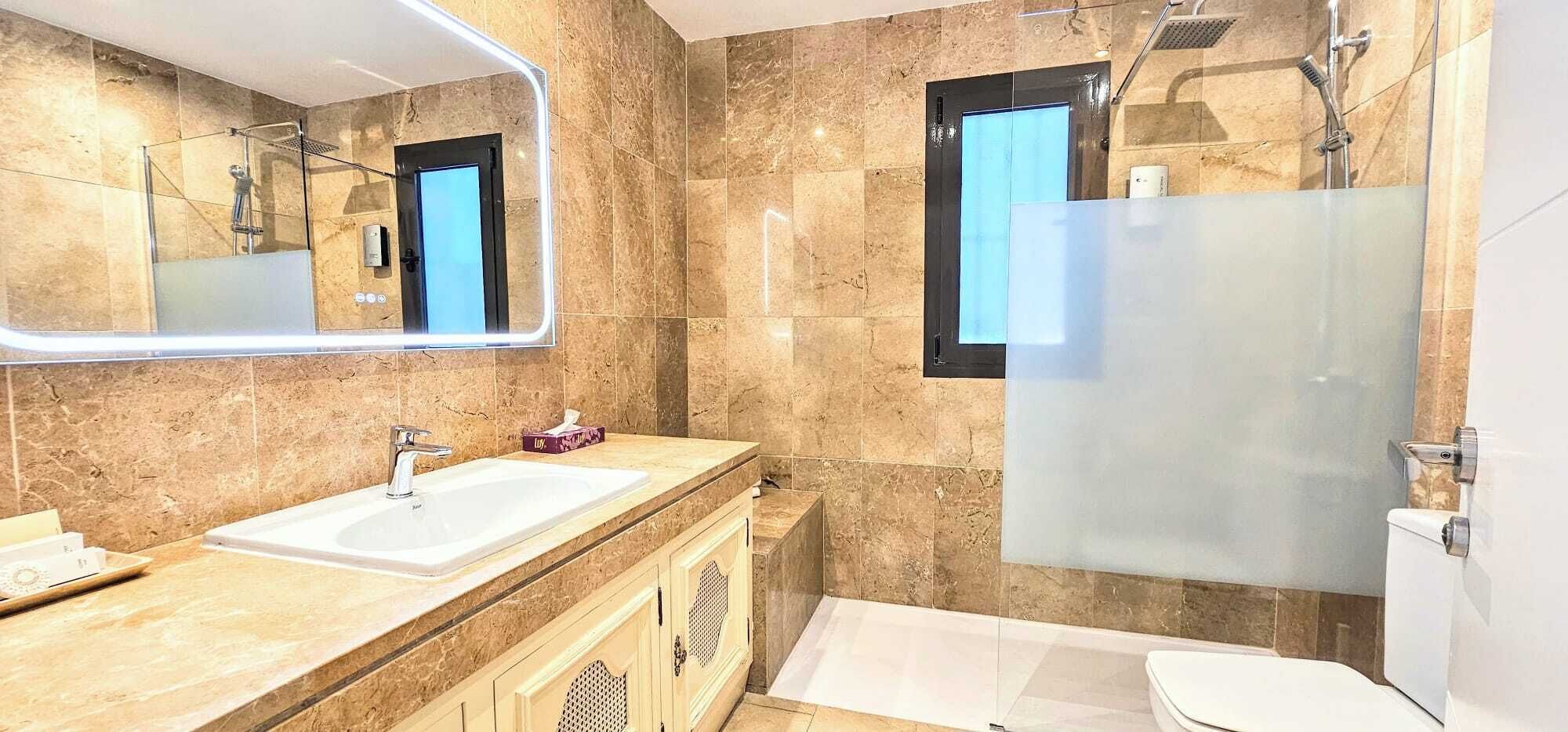 A modern bathroom with a large mirror, two sinks, a shower, and a toilet.