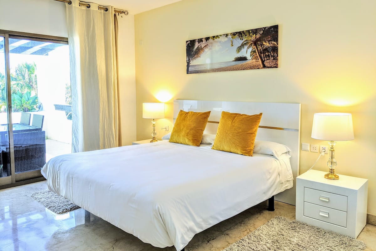 A hotel room with a large bed, two bedside lamps, a painting on the wall, and a view of the outdoors through a sliding door.