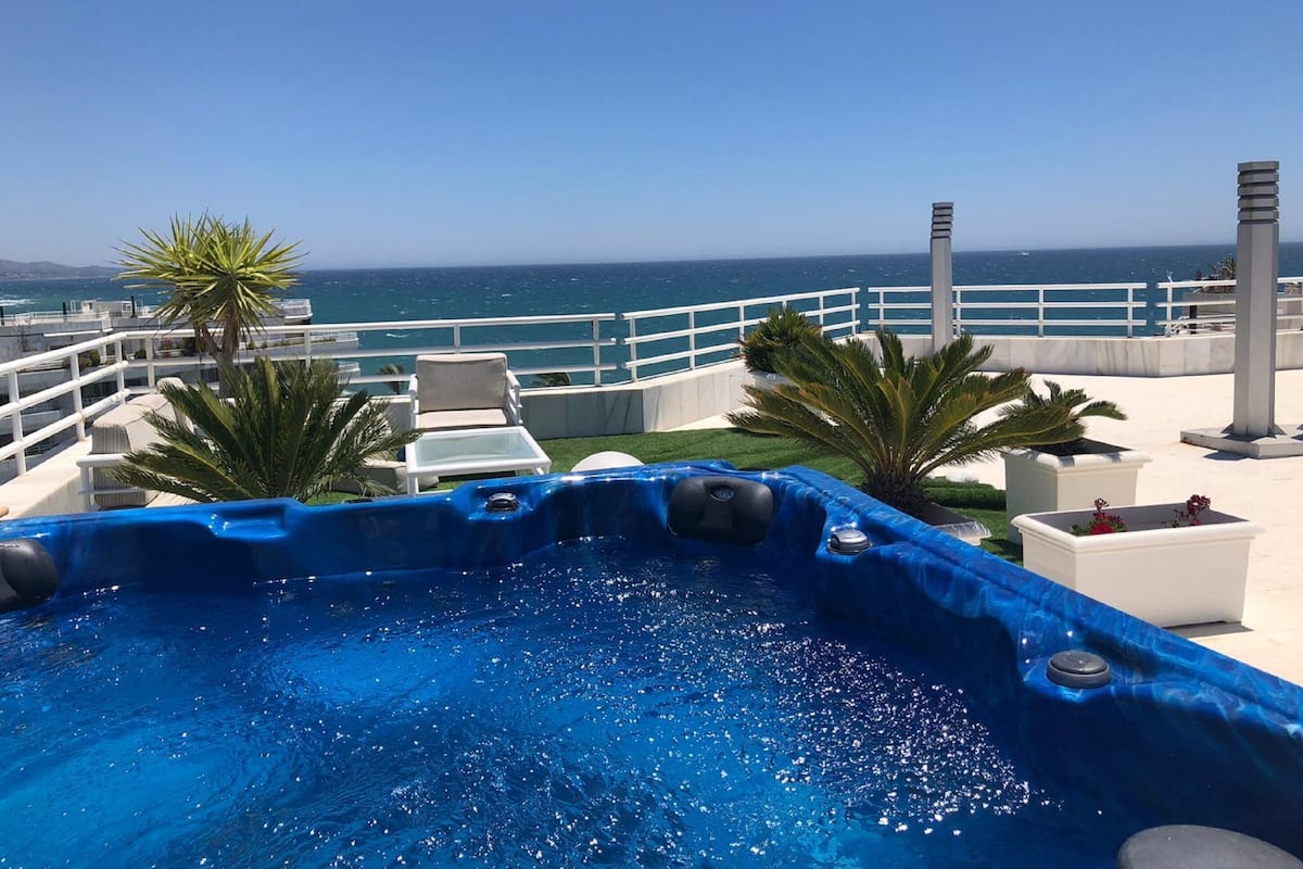 A rooftop pool with a clear view of the ocean, surrounded by lounge chairs and palm trees.