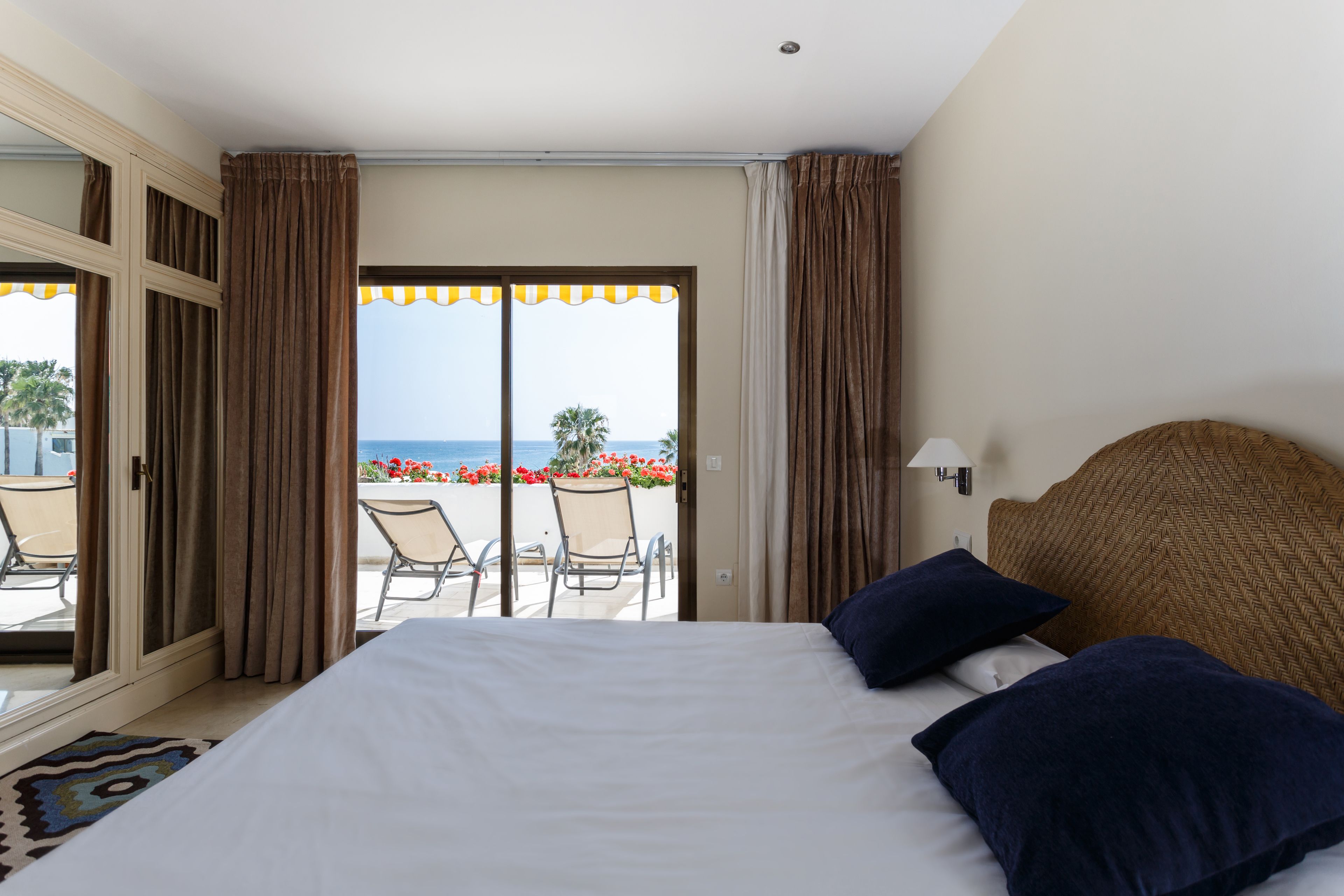 A hotel room with a bed, a view of the sea, and a balcony with chairs and flowers.
