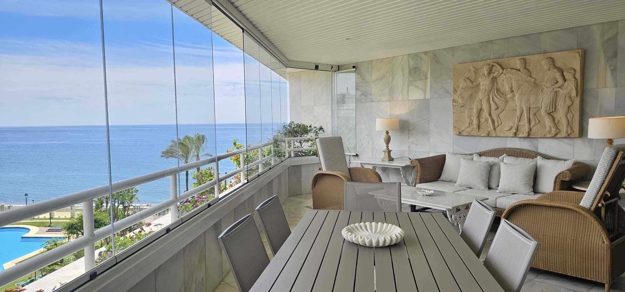 A balcony with a view of the ocean, a dining table, and seating arrangements.