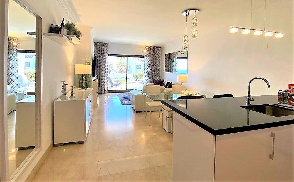 Een moderne keuken met een centraal eiland, een zithoek met een bank en stoelen, en een uitzicht op de omgeving door grote ramen.
