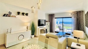 Una sala moderna con una mesa de centro de vidrio, un sofá color beige y un televisor. Hay plantas, una estantería y vistas al mar a través de amplias ventanas.