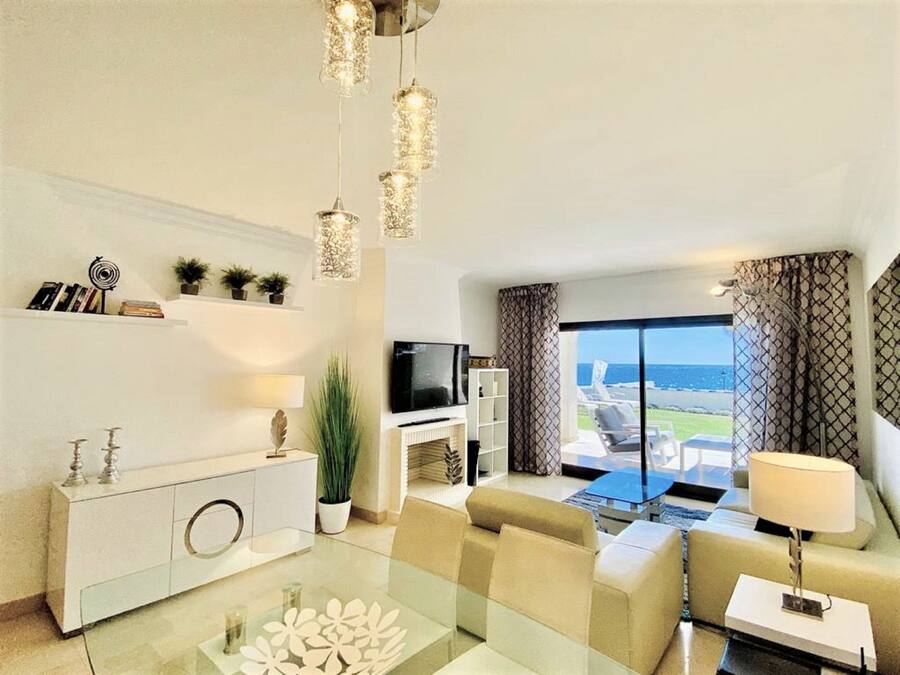 Una sala moderna con una mesa de centro de vidrio, un sofá color beige y un televisor. Hay plantas, una estantería y vistas al mar a través de amplias ventanas.