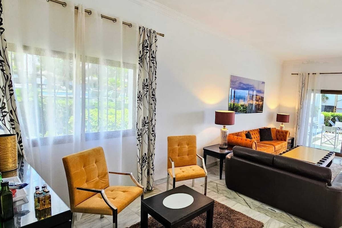 A modern living room with a sofa, two armchairs, a coffee table, and a TV stand. There are two lamps on the coffee table and a painting on the wall.