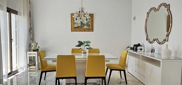 Een eetgedeelte met een witte tafel, vier gele stoelen, een kroonluchter en een spiegel aan de muur.