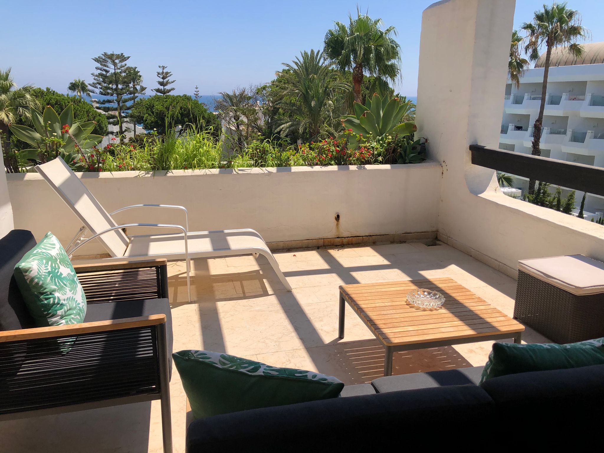 A sunlit outdoor seating area with a sofa, chairs, and a table, surrounded by lush greenery and a view of the sea.