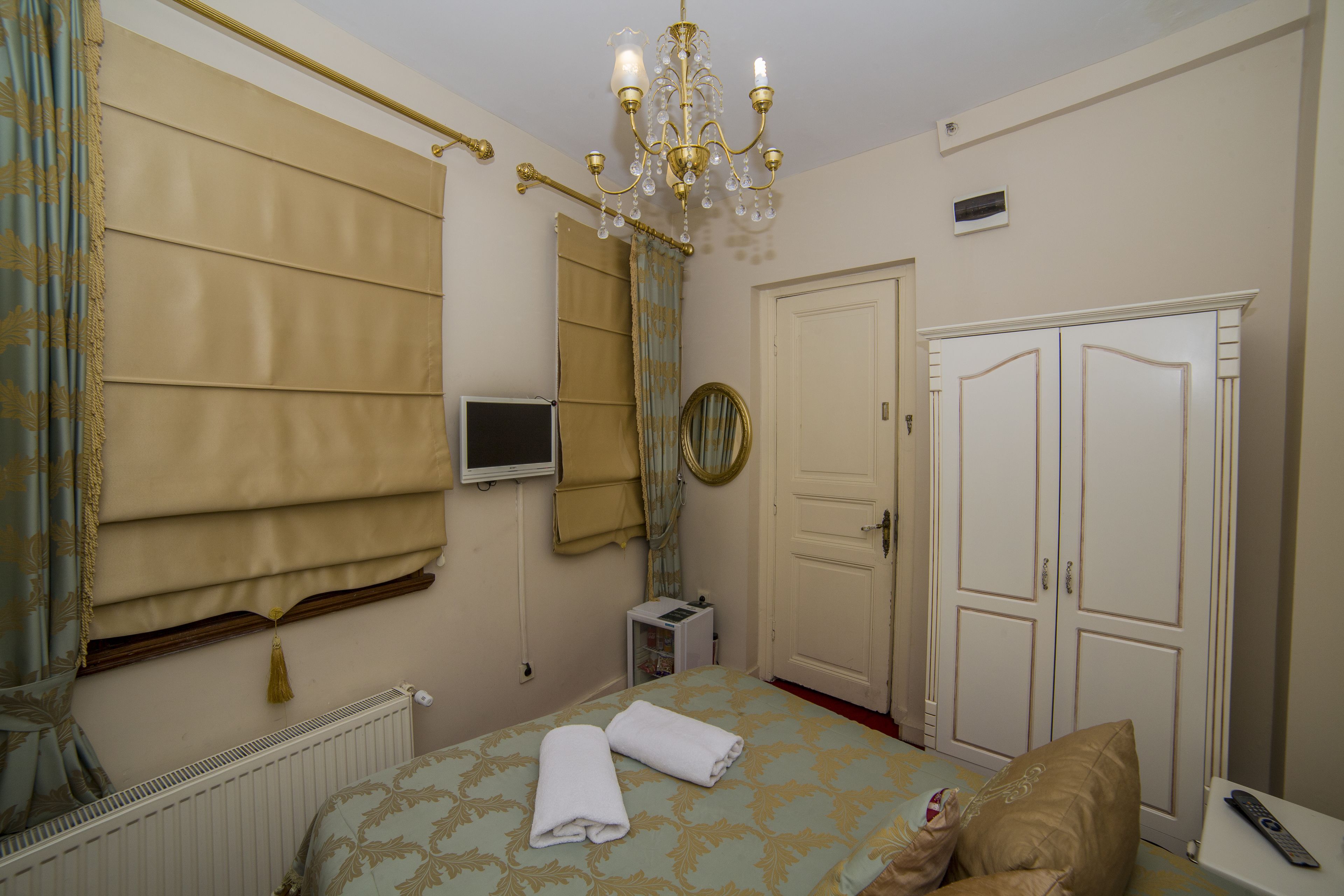 A hotel room with a bed, a chandelier, a wardrobe, a mirror, and a television.