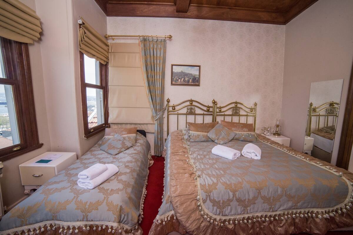 A hotel room with two beds, a red carpet, a window with curtains, a bedside table, a nightstand, a mirror, and a framed picture on the wall.