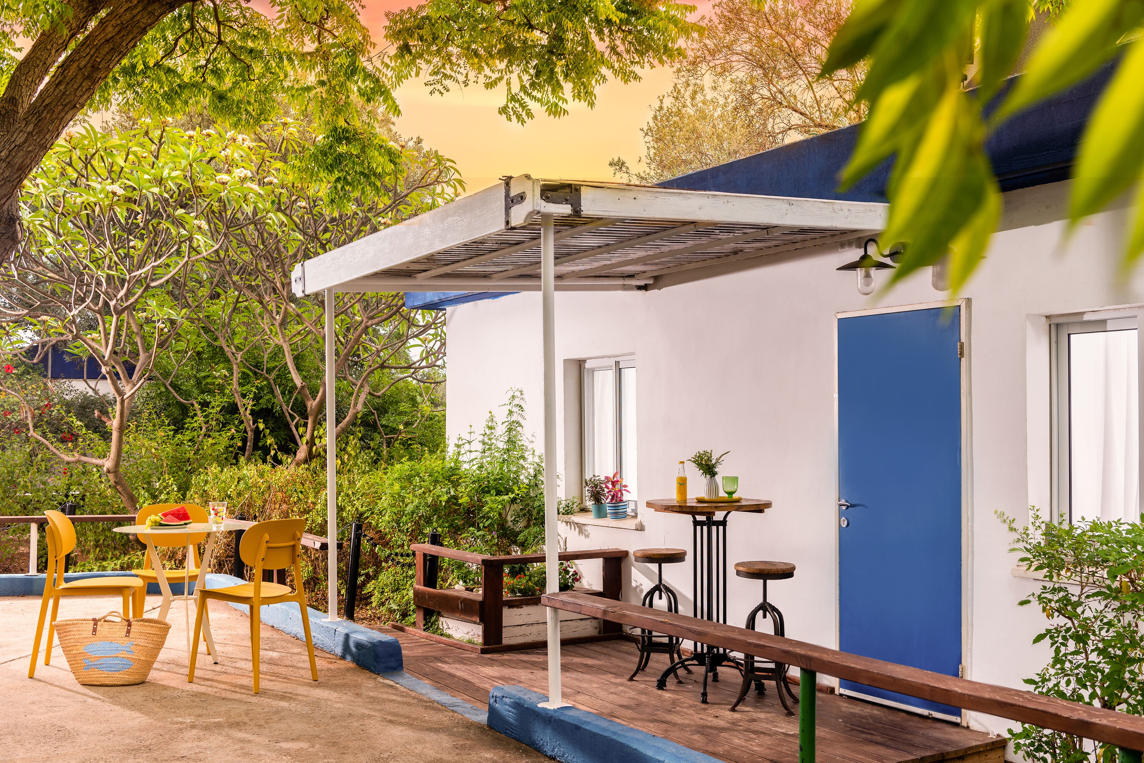 A patio with yellow chairs, a table, and a bench. There is a blue door and a window with a plant on the windowsill.