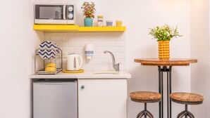 A compact kitchen with a microwave, kettle, and a small refrigerator. There is a sink with a faucet, a shelf with a potted plant, and a round table with stools.