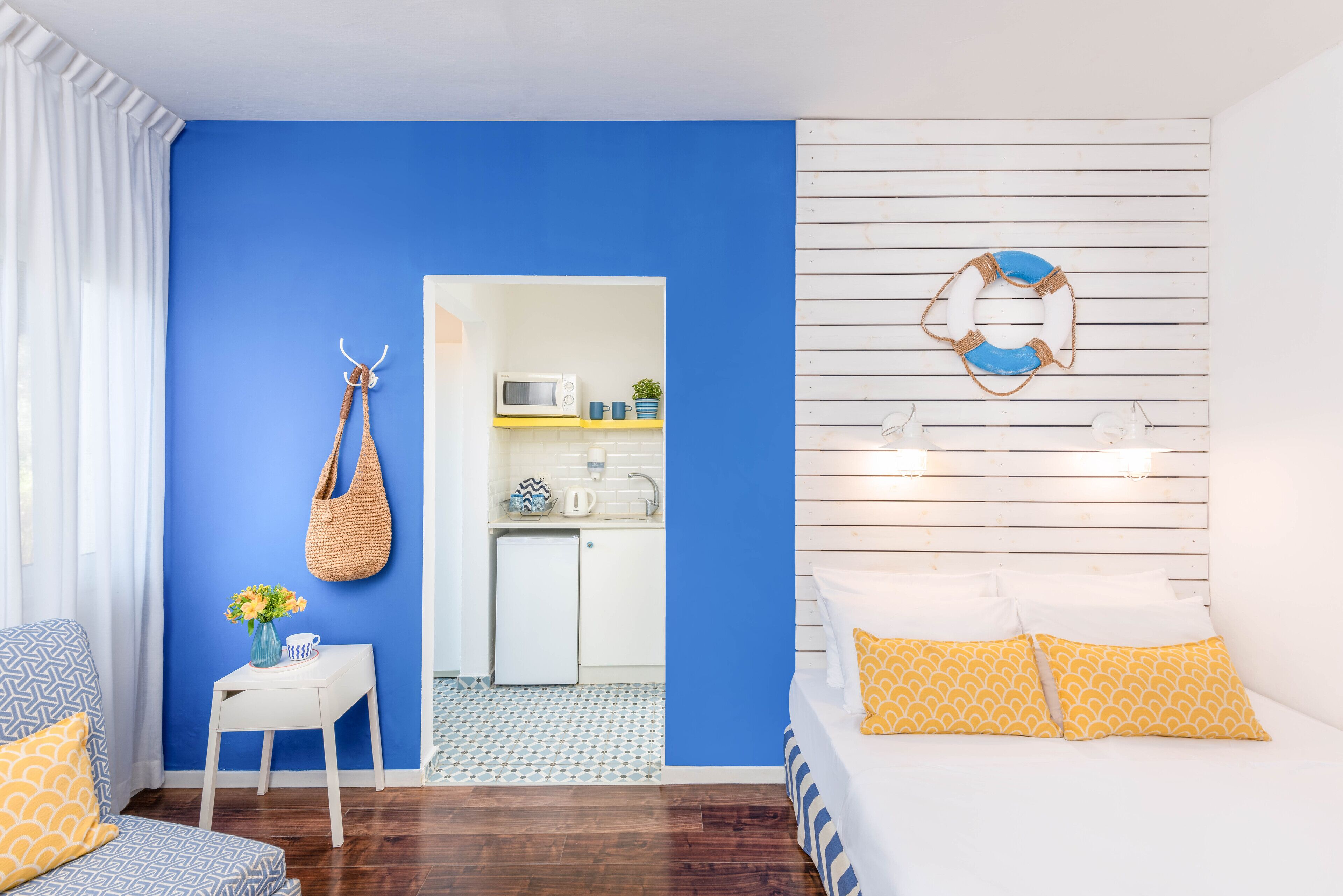 A room with a blue wall, a white bed with yellow pillows, a small white table with a vase, a hanging bag, and a kitchenette in the background.