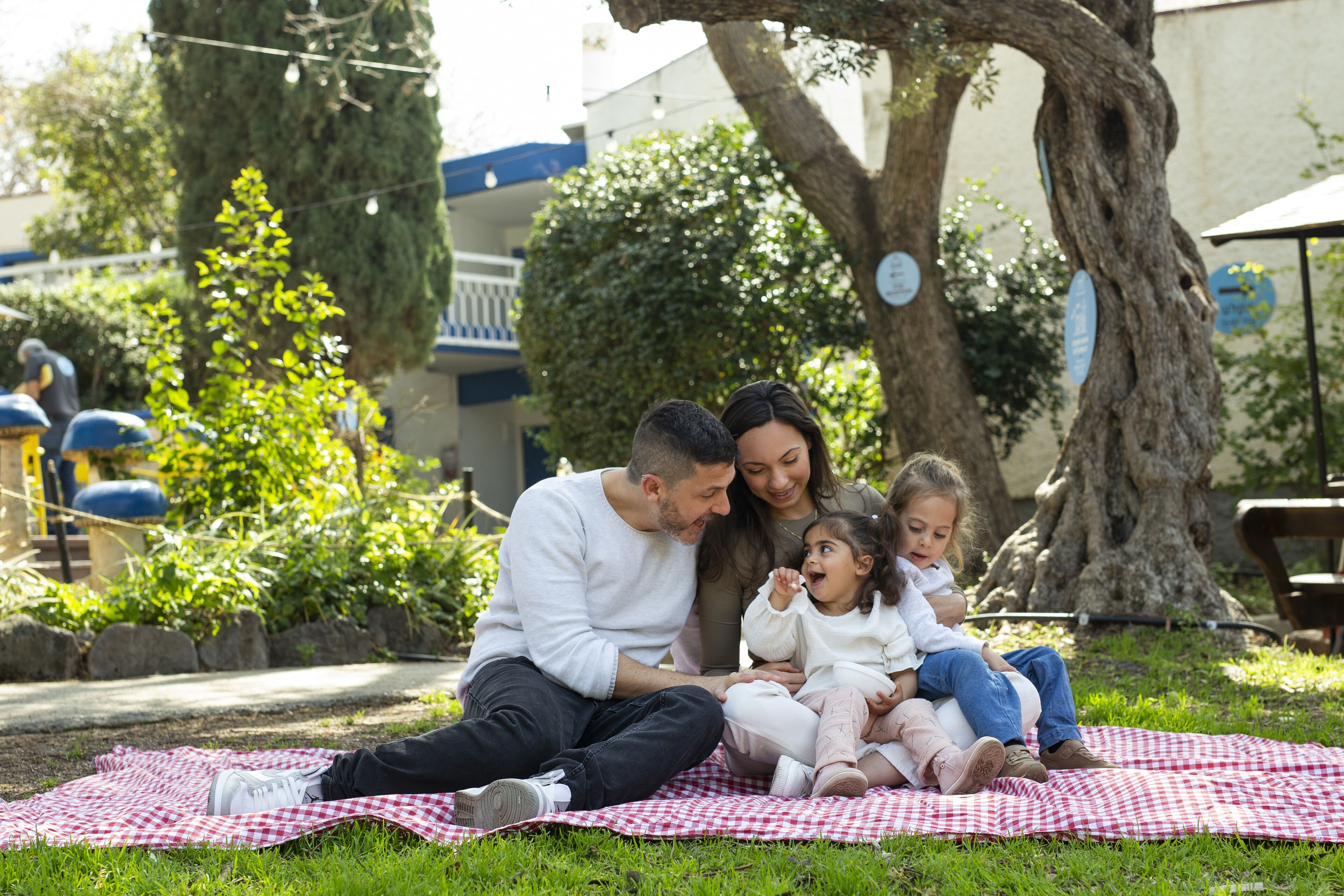 A family sitting on a checkered blanket under a tree in a park.