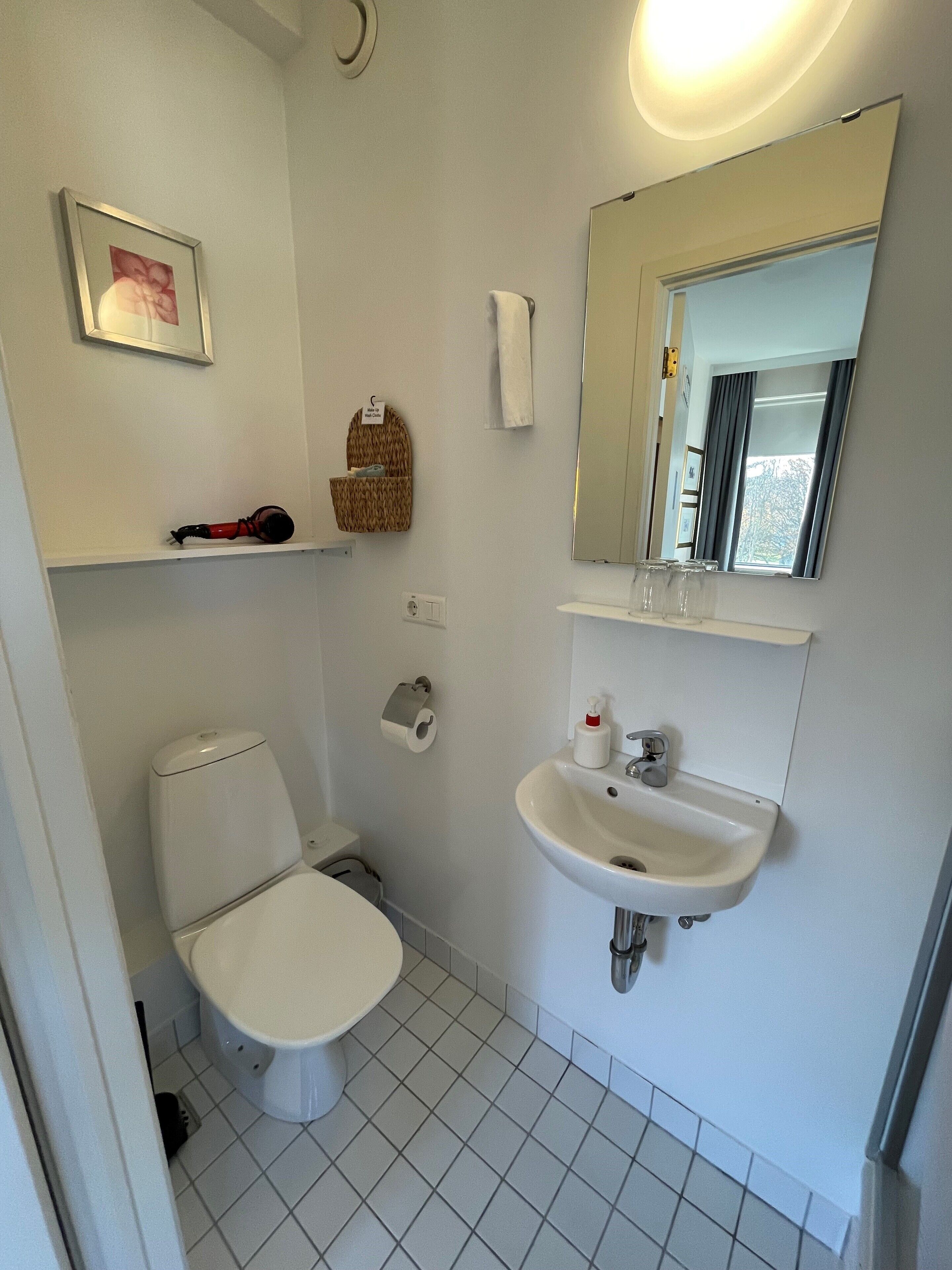 A small, clean bathroom with a toilet, sink, and a mirror above the sink. There is a towel rack and a small shelf with a basket.