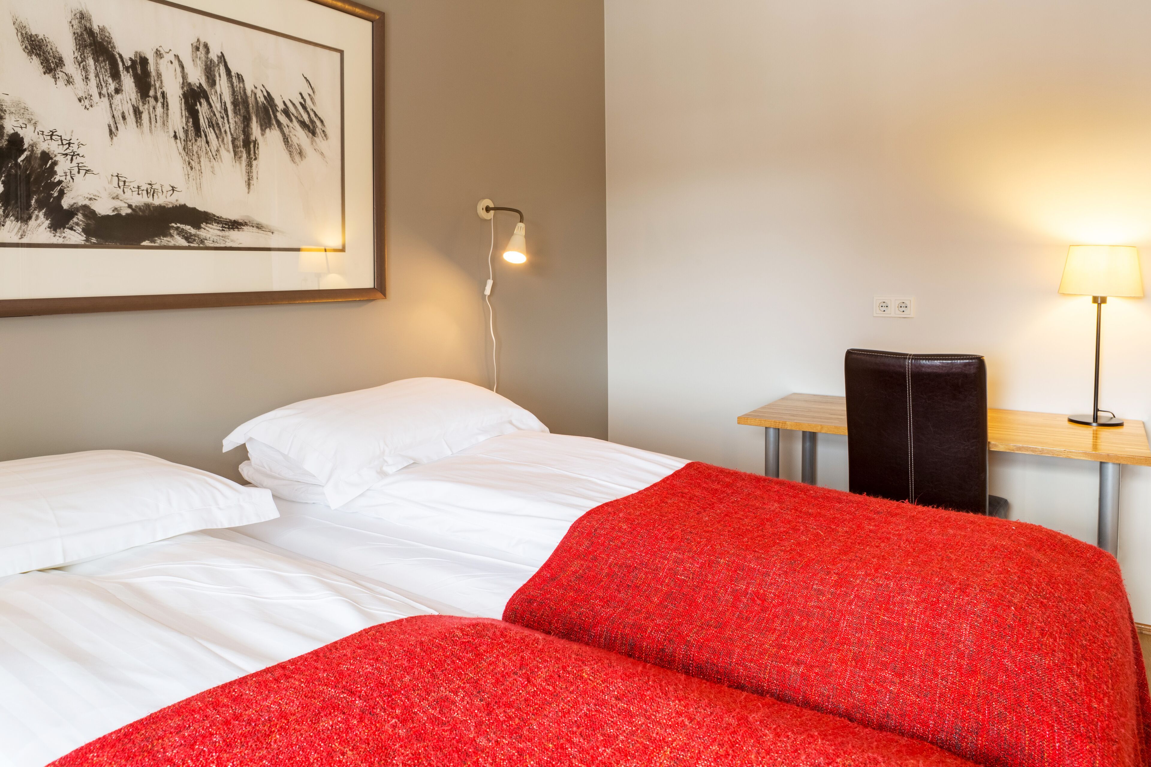 A hotel room with a bed, a desk with a lamp, and a framed artwork on the wall.