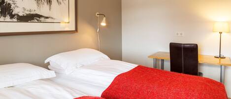 A hotel room with a bed, a desk with a lamp, and a framed artwork on the wall.
