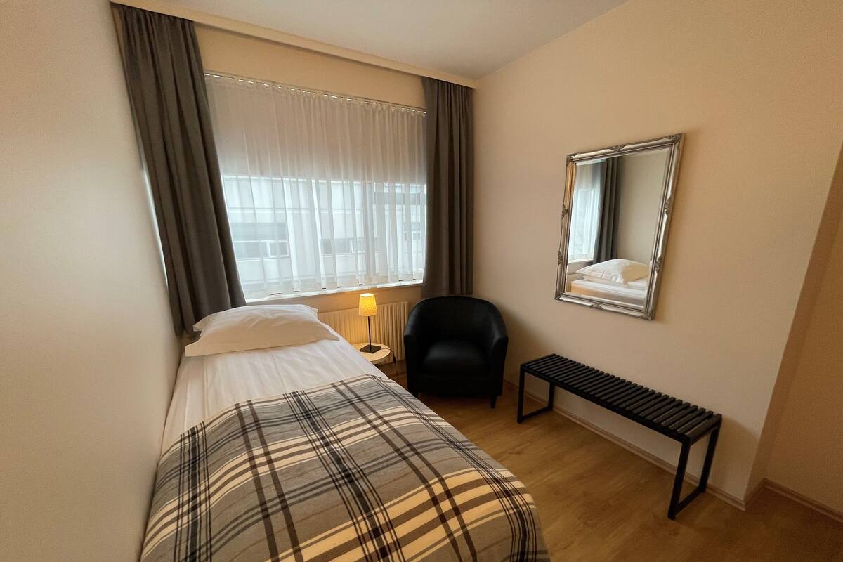 Quarto de hotel com cama, poltrona, mesinha, abajur e espelho na parede.
