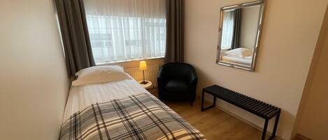 A hotel room with a bed, a chair, a small table, a lamp, and a mirror on the wall.